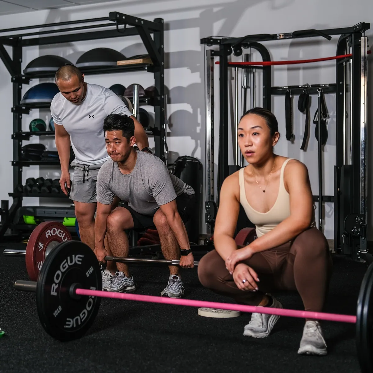 A Formula Fitness coach and two clients in a squat position around a barbell during a coached training session